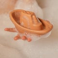 Unrecognizable child hold cute orange small silicone toy boat with holes on top with hand in bubble bath. Square frame. Royalty Free Stock Photo