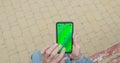 Unrecognizable elderly mans hand uses smartphone. Green screen, swipe, touch, top view. Bench, paving slabs, outdoors.