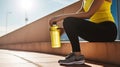 Unrecognizable female athlete sitting with water bottle on concrete platform in bright sunlight while taking break from training.
