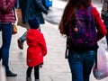 An unrecognizable girl in a red coat and blue hat is standing next to her mother on the street and holding a doll in her hands.