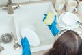 Unrecognizable little preteen girl wearing big blue rubber gloves, holding soapy sponge, washing white plate in kitchen. Royalty Free Stock Photo