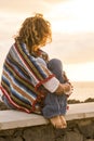 Unrecognizable lonely woman sit down on a wall looking and enjoying the sunset on the sea - coloured trendy mexican poncho style Royalty Free Stock Photo