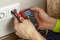 An unrecognizable male technician measures the voltage at the outlet with a digital multimeter. There are only hands in the frame