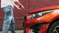 unrecognizable man in jeans and white shirt opens hood of orange car parked near brick building. concept of breakage