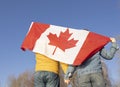 unrecognizable man and woman, holding hands, stand with a large Canadian flag behind their back against the sky