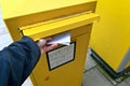 Unrecognizable person putting an envelope into the mail slot of a yellow letterbox.