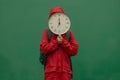 Unrecognizable person in red outfit hiding face behind big round clock with white dial. A human in red suit with big