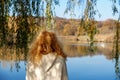 Unrecognizable redheaded woman with curly hair from behind under a willow tree in autumn looking at a lake with two swans Royalty Free Stock Photo