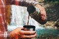 Unrecognizable Traveler Pouring Tea Or Coffee To Cup From Thermos On Waterfall Background. Tourism And Travel Concept