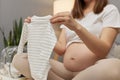 Unrecognizable unknown pregnant woman preparing baby clothes for maternity hospital in home interior admiring tiny striped attire Royalty Free Stock Photo
