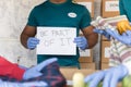 Unrecognizable volunteer with medical face mask showing Be Part of It sign board while other busy packing donations - concept of