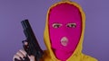 Unrecognizable woman in pink balaclava with gun on purple background. Dangerous criminal in mask with pistol looking at