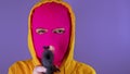 Unrecognizable woman in pink balaclava with gun on purple background. Dangerous female in mask takes aim at camera and