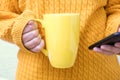 Unrecognizable woman in yellow autumn knitted sweater holding a big cup, mug of coffee and using smartphone, fall Royalty Free Stock Photo