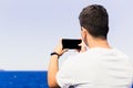 Unrecognizable young man with his back and brown hair, taking a horizontal photo of the beach with a smartphone, generic