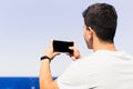 Unrecognizable young man with his back and brown hair, taking a horizontal photo of the beach with a smartphone, generic