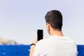Unrecognizable young man with his back and brown hair, taking a vertical photo of the beach with a smartphone, generic photography