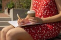 Unrecognizable Young woman in red dress drinking coffee from craft paper cup Writing Gratitude Journal on wooden bench