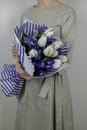 Unrecognizible woman in beige dress holding bouquet of white tulips and violet iris in striped lilac wrapping paper.