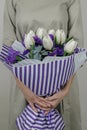 Unrecognizible woman in beige dress holding bouquet of white tulips and violet iris in striped lilac wrapping paper.