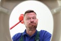 Unshaven man looks into toilet bowl holding red plunger Royalty Free Stock Photo