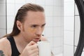 Unshaven man with patches under his eyes and cup of coffee looks at himself in bathroom mirror Royalty Free Stock Photo