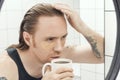 Unshaven man with patches under his eyes and cup of coffee looks at himself in the bathroom mirror. Male with morning hangover Royalty Free Stock Photo