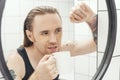 Unshaven man with patches under his eyes and cup of coffee looks at himself in bathroom mirror Royalty Free Stock Photo