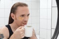 Unshaven man with patches under his eyes and cup of coffee looks at himself in bathroom mirror Royalty Free Stock Photo