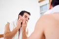Unshaven man squeezing pimple on face with white towel on neck standing near mirror