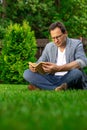 Unshaven 40s man in glasses sits on grass reading book outdoors on summer day Royalty Free Stock Photo