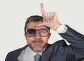 Unshaven and werid businessman in weird broken nerdy glasses and stupid smile doing loser sign with L fingers signal on forehead