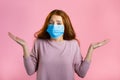 Unsure woman in medical mask shrugs her arms, makes gesture of I don`t know, care, can`t help anything . Girl on pink