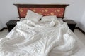 Untidy / unmade bed with two messy pillows and a white crumpled blanket in a hotel.