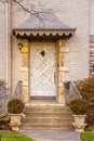 Unusual rustic wooden door surrounded by rock trim in light colored upscale brick house with awning on winter day with all bushes