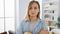 Unwavering young blonde business woman, a picture of serious concentration, stands with arms crossed in the vibrant office