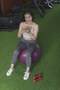 An upbeat and cheerful woman smiling at the camera while holding her phone and sitting on a swiss ball inside a gym. Updating her Royalty Free Stock Photo