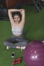 An upbeat and cheerful woman stretching her arms while sitting cross-legged on the faux grass at the gym, with a swiss ball beside