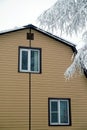 Upper floor with plastic window  in country house wall covered with beige siding and brown metal roof with snow surface and tree b Royalty Free Stock Photo