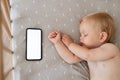 Upper view of baby napping in his bed next to phone with blank white screen