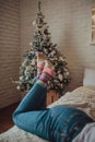 Upright female legs in skinny jeans and bright knitted socks on a bright bed against the background of the Christmas tree