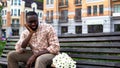 Upset afro-american man sitting lonely on city bench with bouquet, failed date