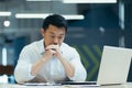 Upset Asian male businessman sitting in the office at the table, thoughtfully leaning his head on his hands. Financial Royalty Free Stock Photo