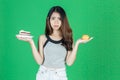 Upset Asian woman choosing between orange fruit or unhealthy cake on hands over green  background. Healthy lifestyle and Royalty Free Stock Photo