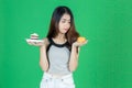 Upset Asian woman choosing between orange fruit or unhealthy cake on hands over green isolated background. Healthy lifestyle and Royalty Free Stock Photo