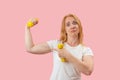 Upset attractive young woman with dumbbells. Grueling workout, Sporty red hair girl. Pink pastel background