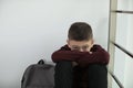 Upset boy with backpack sitting near wall indoors.