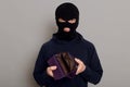 Upset burglar dressed in black turtleneck and balaclava holding empty wallet, feeling frustrated, looking at camera with pouting