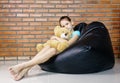 Upset caucasian teen girl sitting in black bean bag chair holding soft teddy bear toy against brick wall. Casual outfit. Child Royalty Free Stock Photo