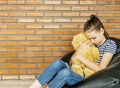Upset caucasian teen girl sitting in black bean bag chair hug big brown teddy bear toy against brick wall. Casual outfit. Sadness